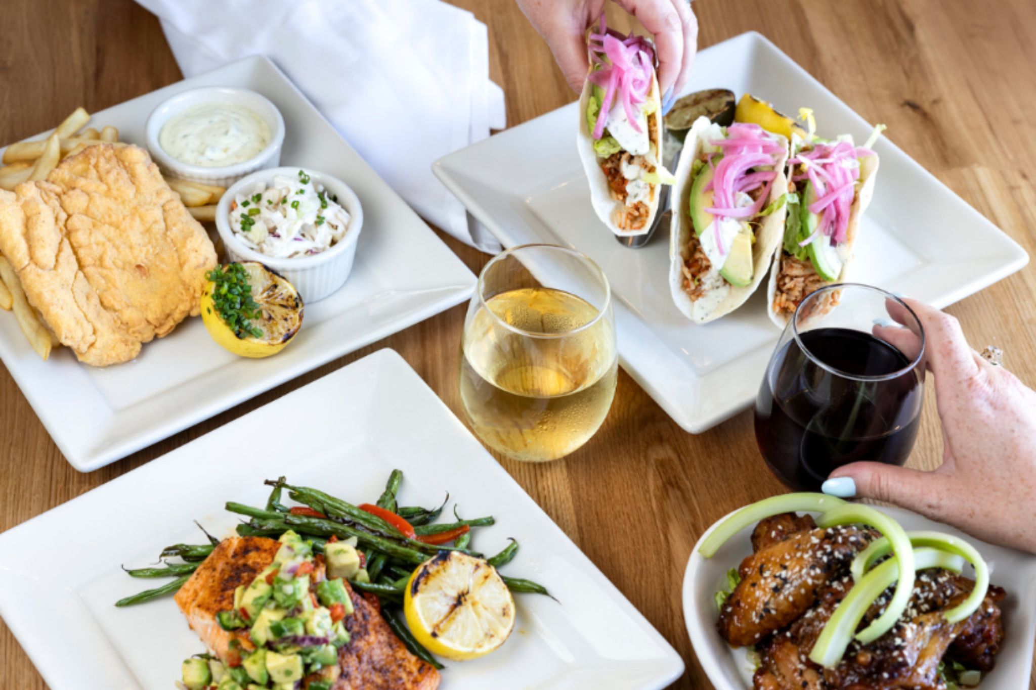 Plates of fish tacos, grilled salmon, chicken wings, and drinks on a wooden table.