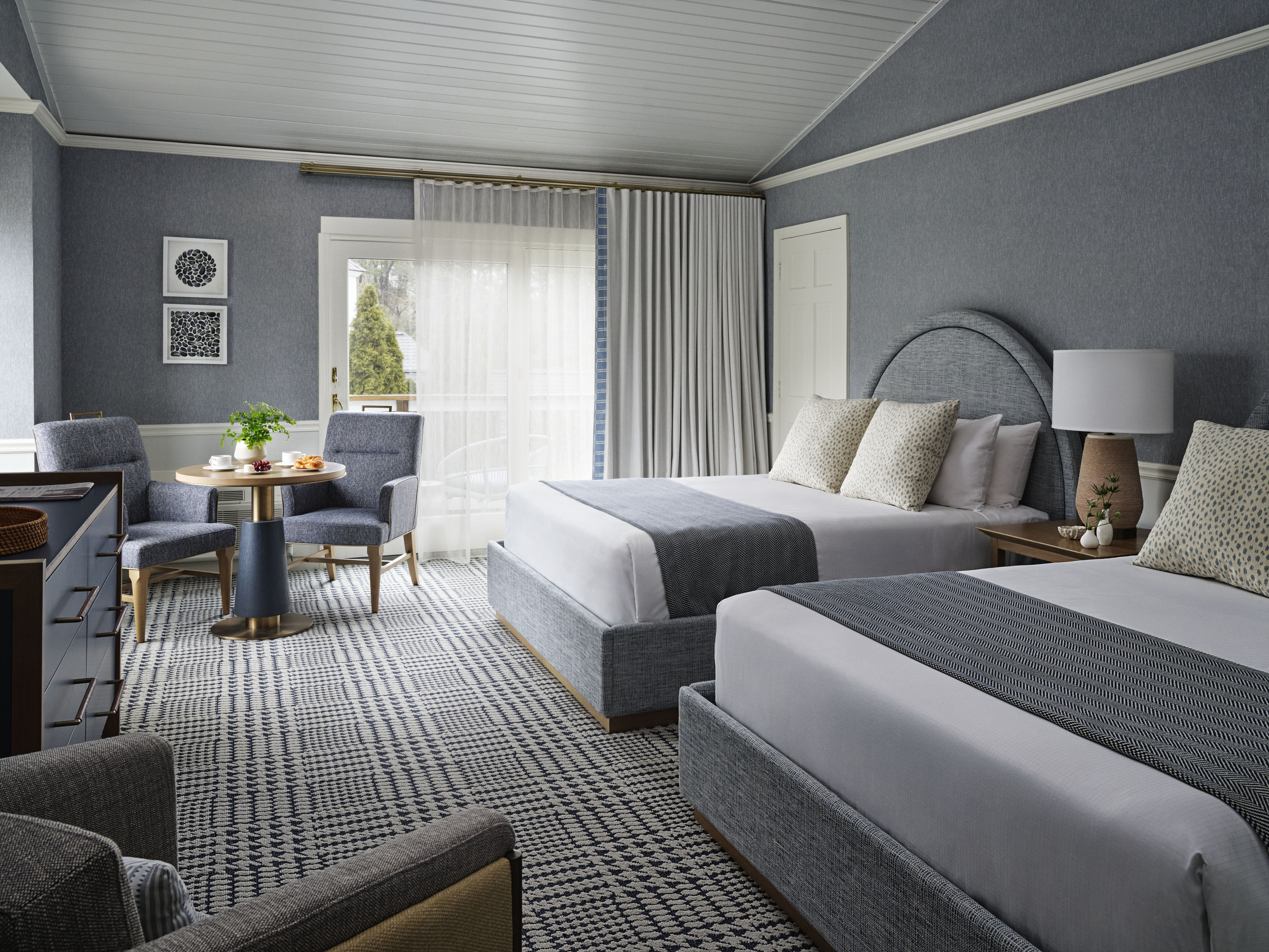 Two double beds in a modern hotel room with gray decor and a small dining area.