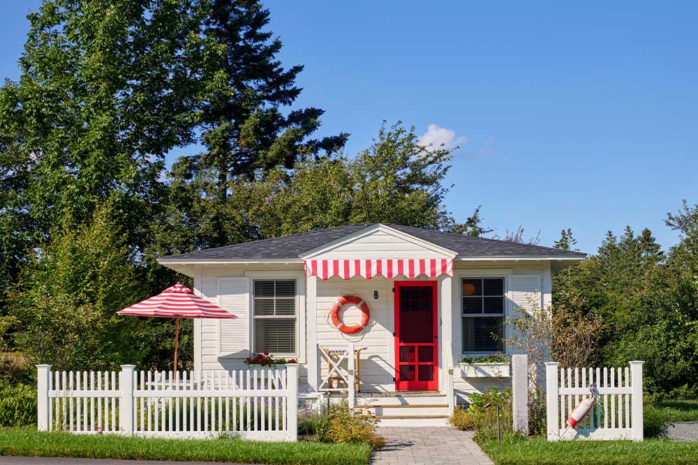 Small white cottage with red door, picket fence, and striped awning.