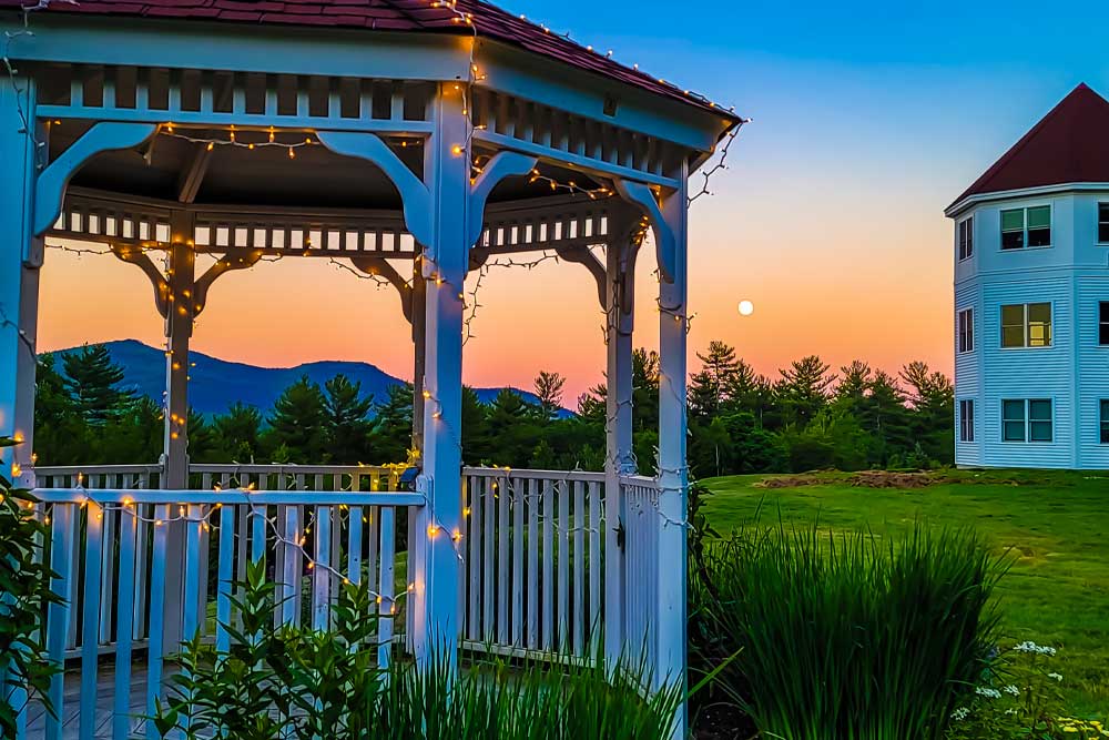 Gazebo with string lights at sunset, mountains and house in background.