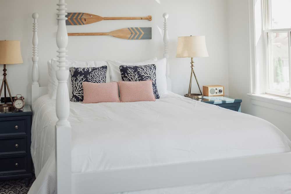 White bedroom with a four-poster bed, nautical decor, and sunlight through a window.