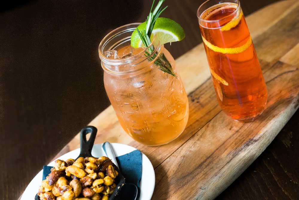 Two cocktails with garnishes on a wooden board, next to a bowl of mixed nuts.