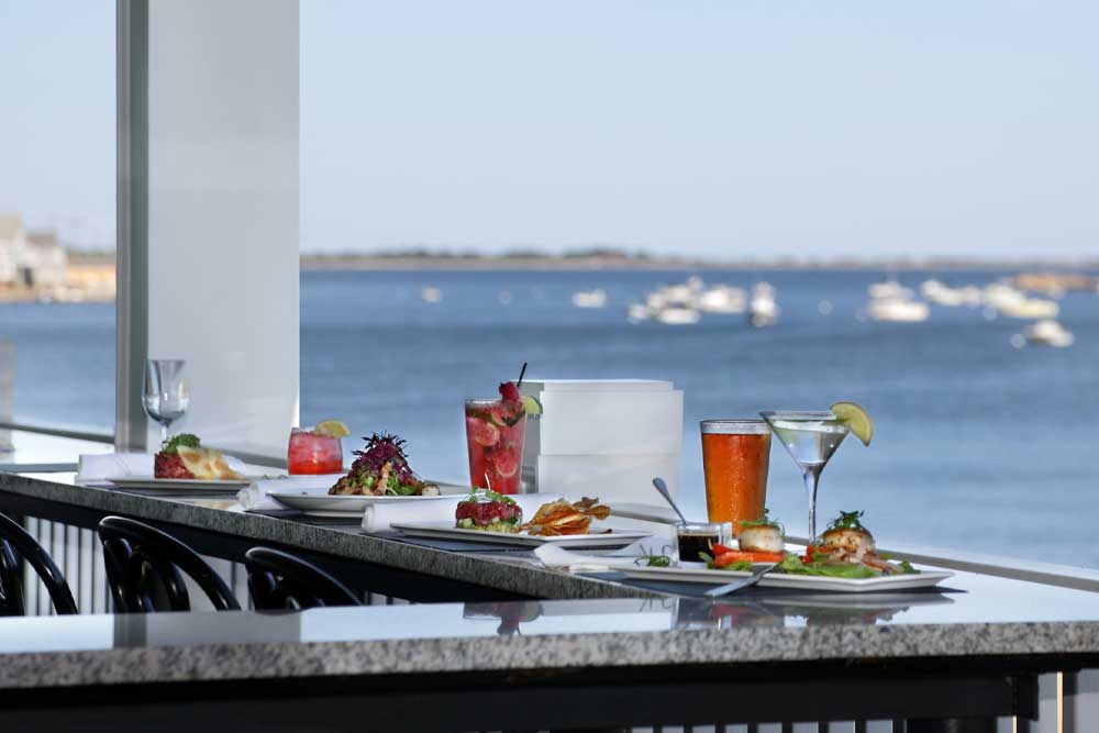 Dining table with drinks and food overlooking a calm waterfront.