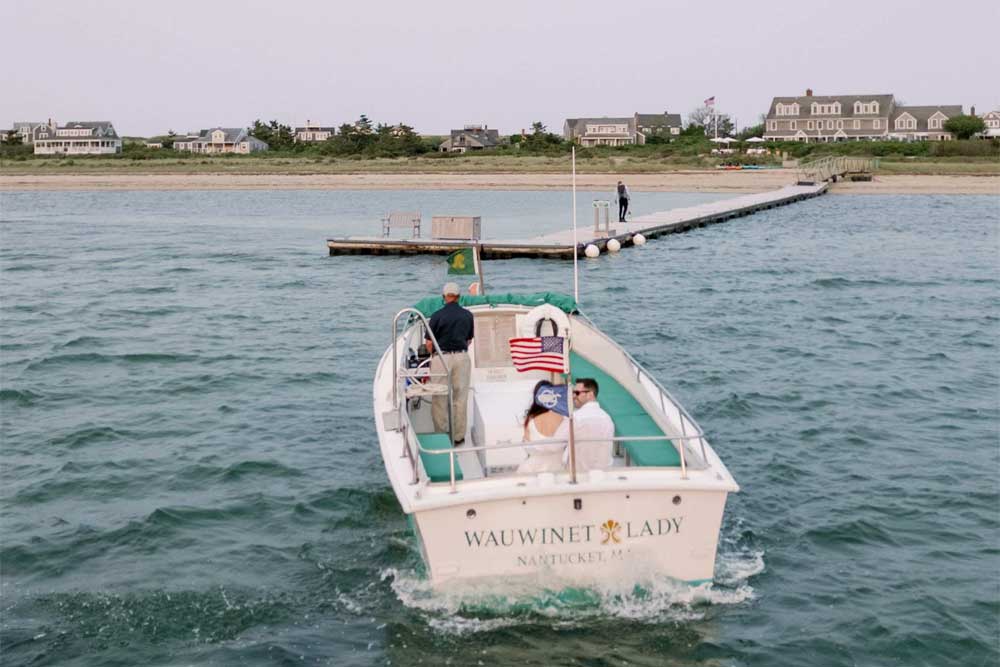 Small boat with people approaching a dock, calm water, houses in the background.