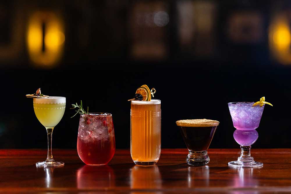 Five colorful cocktails on a bar counter in dim lighting.