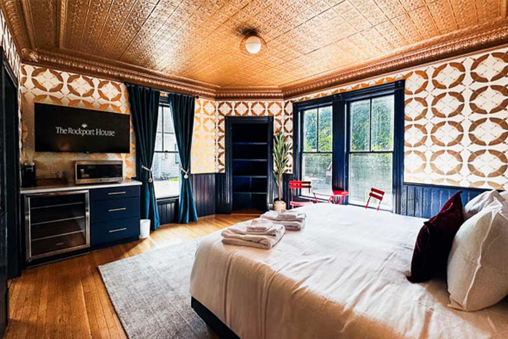 Hotel room with a bed, TV, kitchenette, and large windows overlooking greenery.