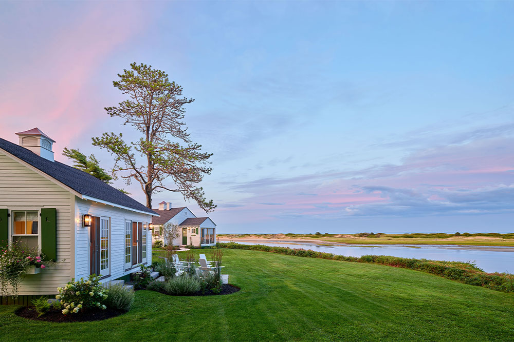 Cozy white cottages by a peaceful lake, under a pink and blue sunset sky.