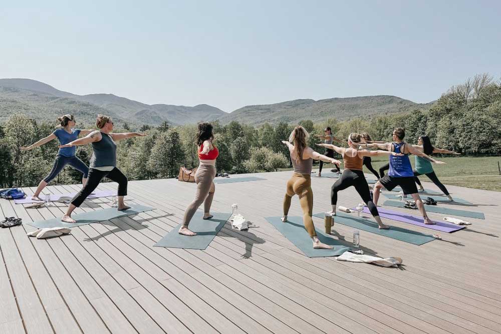 People doing yoga outdoors on a sunny deck, mountains in the background.