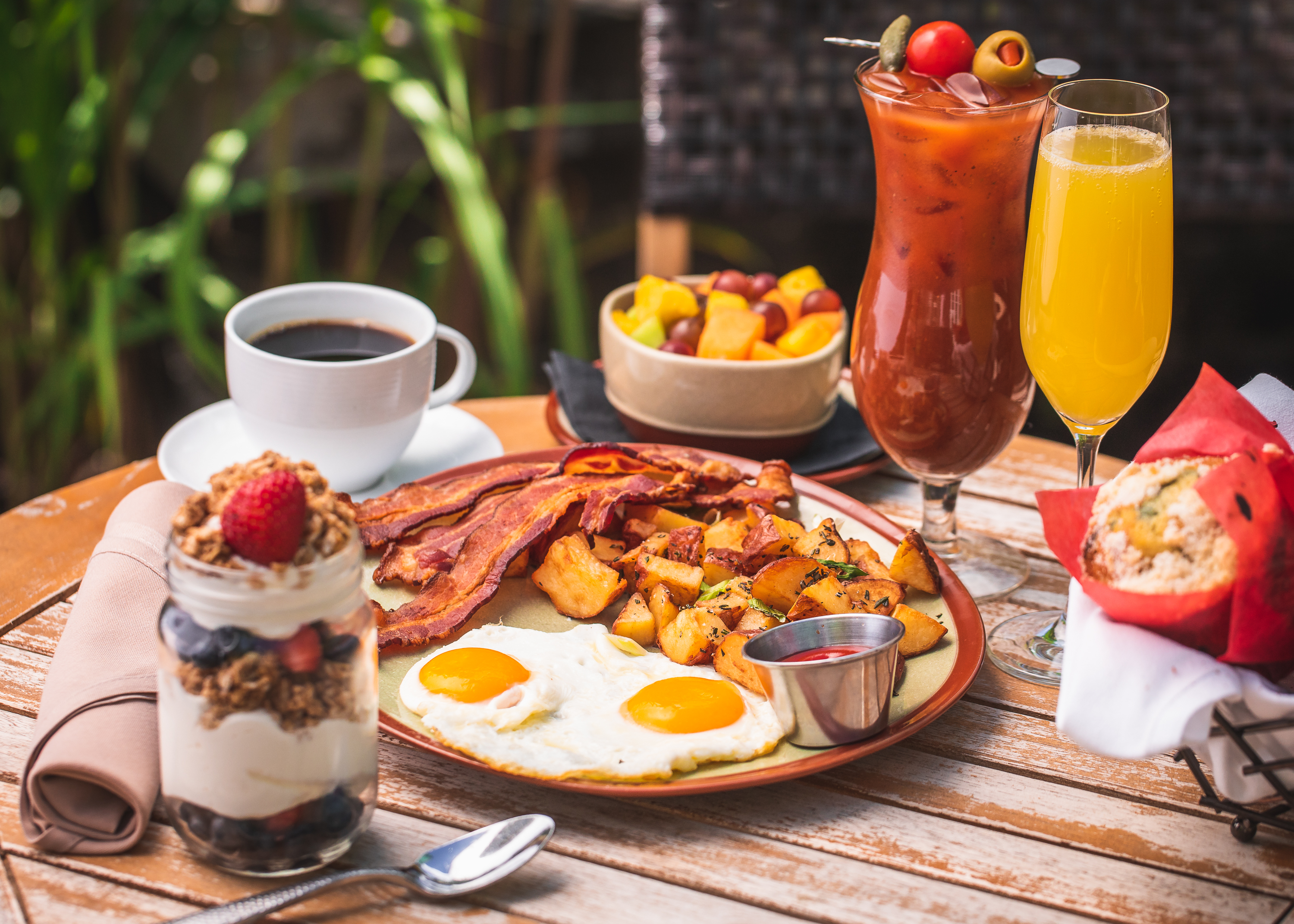 Breakfast spread with eggs, bacon, potatoes, fruit, yogurt parfait, coffee, and cocktails.