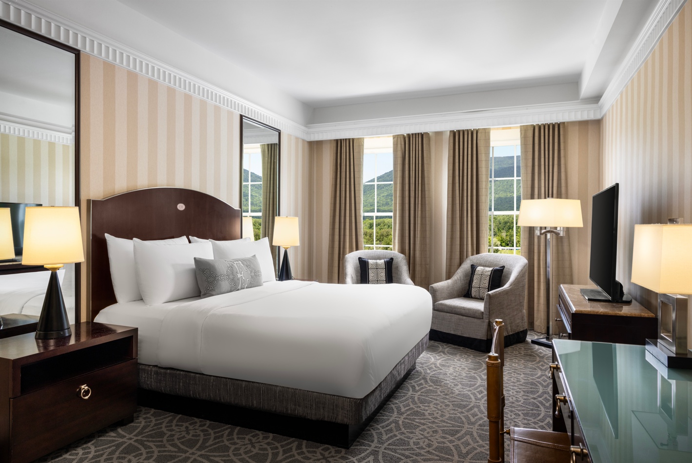 Elegant hotel room with a large bed, armchair, desk, and warm lamps.