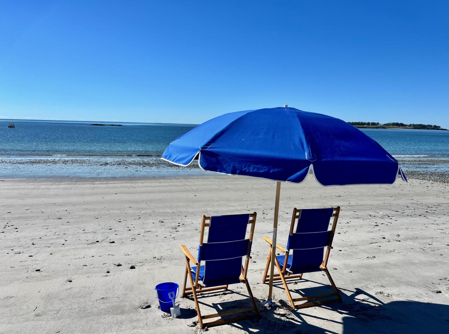 Two empty blue chairs under an umbrella on a sandy beach.