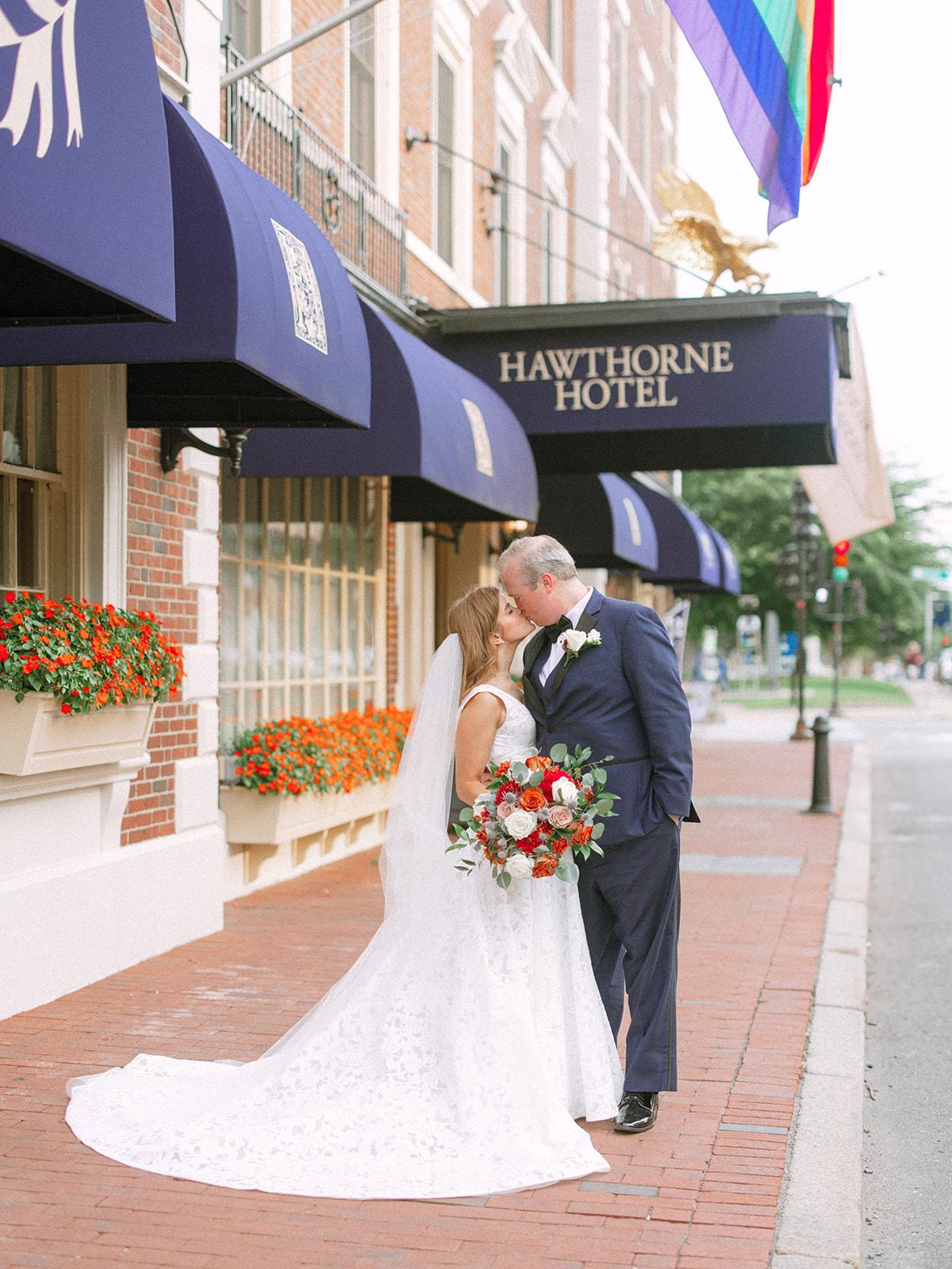 Bride and groom kissing outside a hotel, with a colorful flag above.
