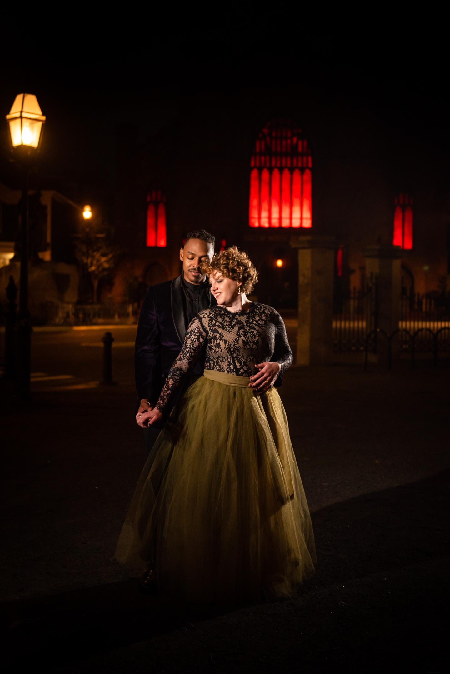 Couple in elegant attire walking at night, red lights in the background.