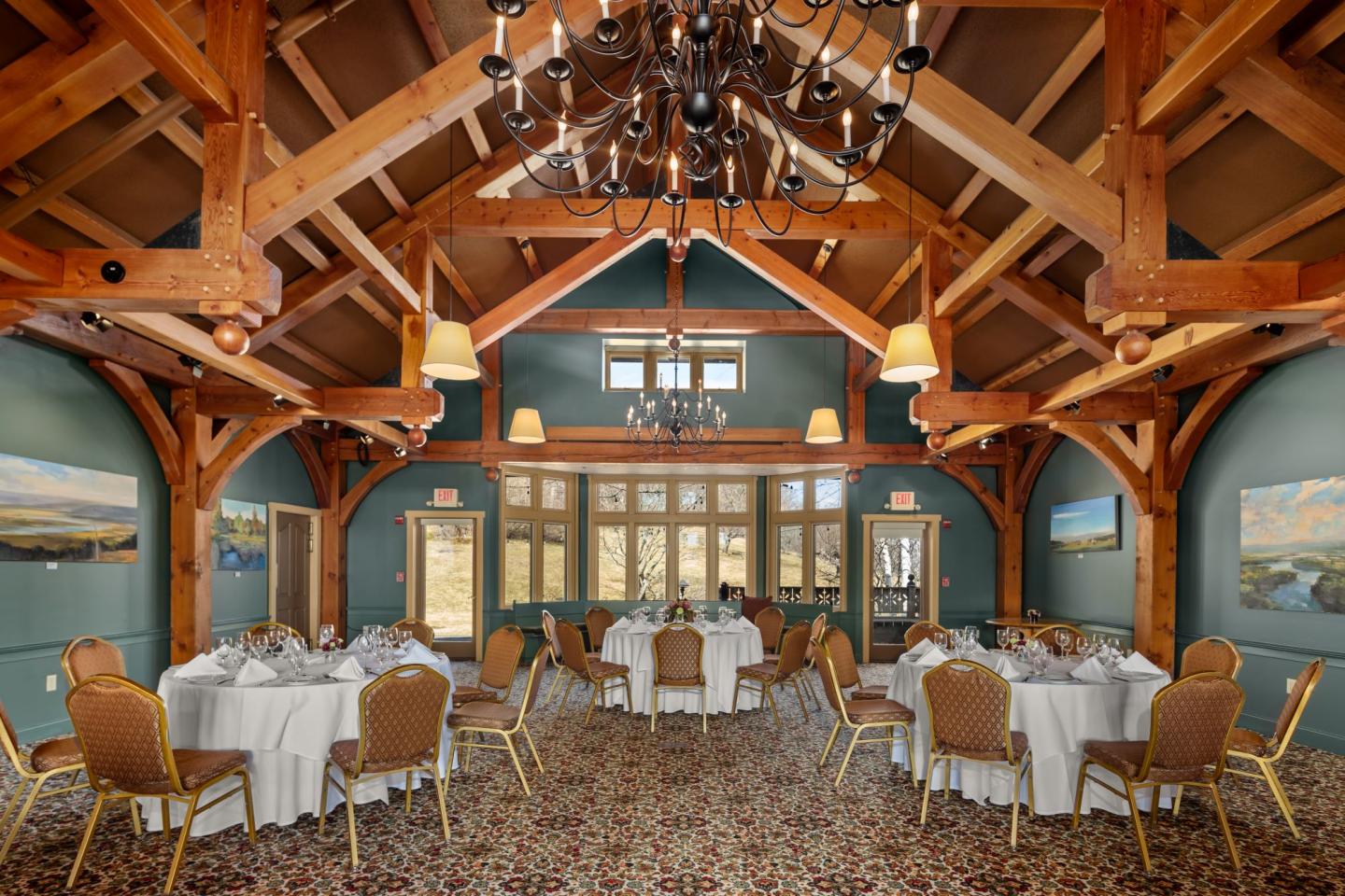 Rustic banquet hall with wooden beams, round tables, white linens, and chandeliers.