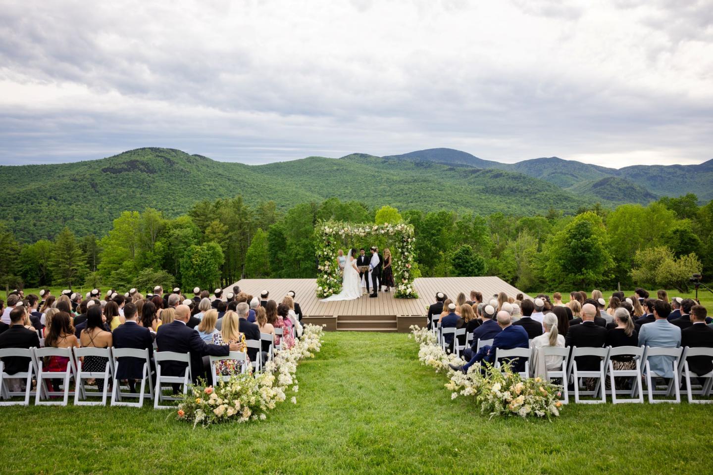 Outdoor wedding ceremony with mountains and guests seated on white chairs.