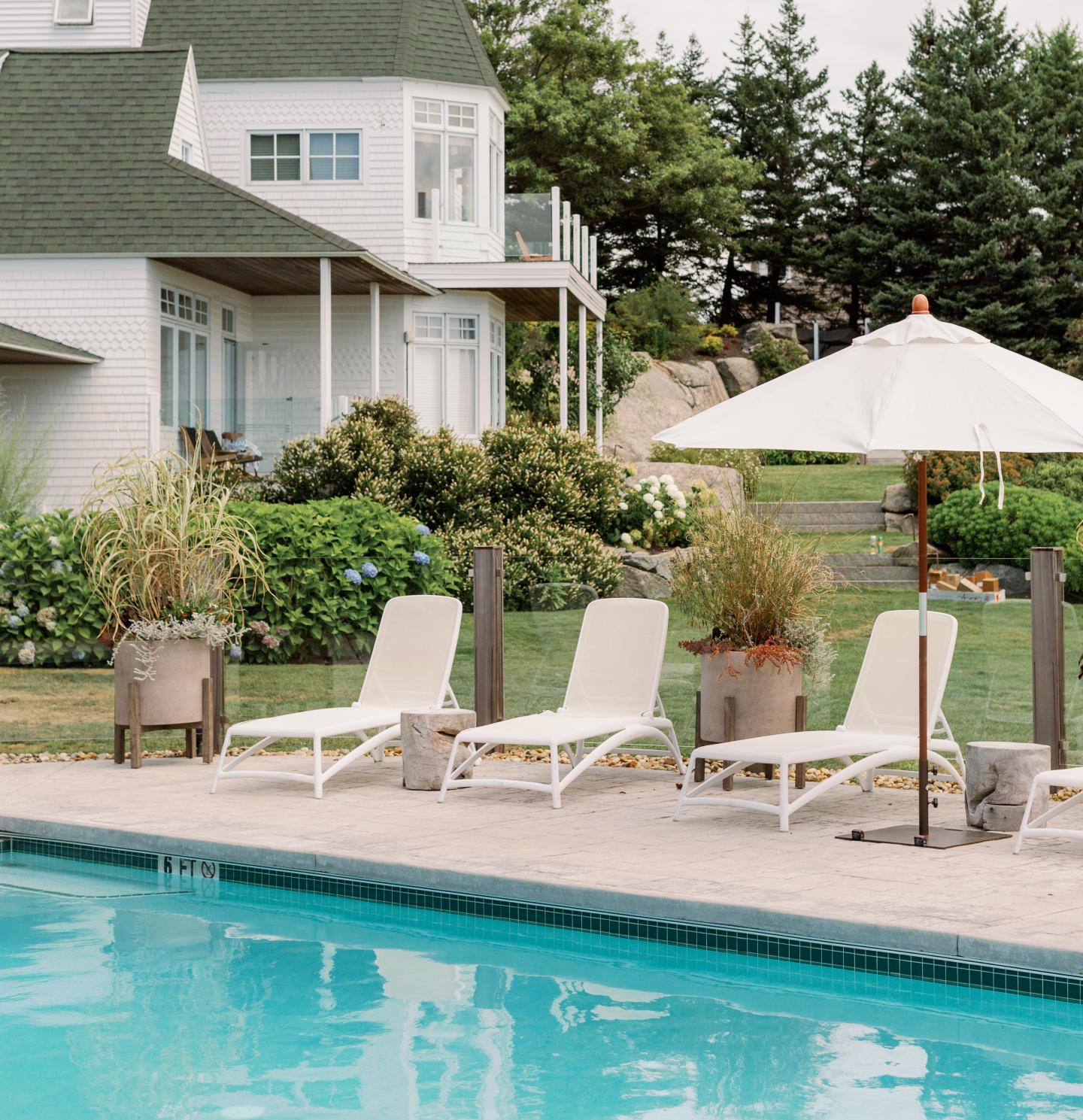 Poolside with lounge chairs and umbrella, house and greenery in the background.