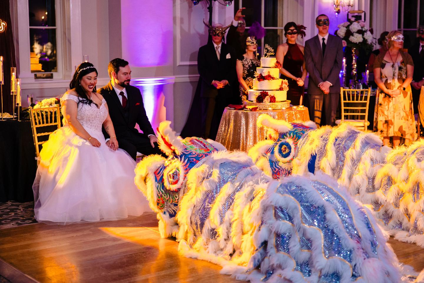 Bride and groom watching a lion dance, wedding guests and cake in background.