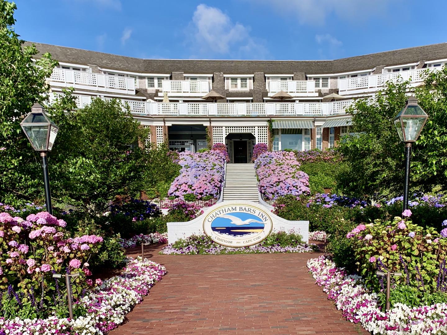 Hotel entrance with colorful floral garden and a white sign displaying a logo.