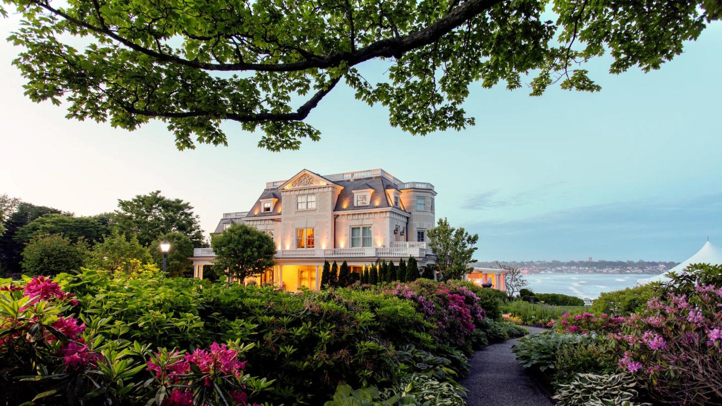 Grand house amidst lush garden overlooking the water at sunset.