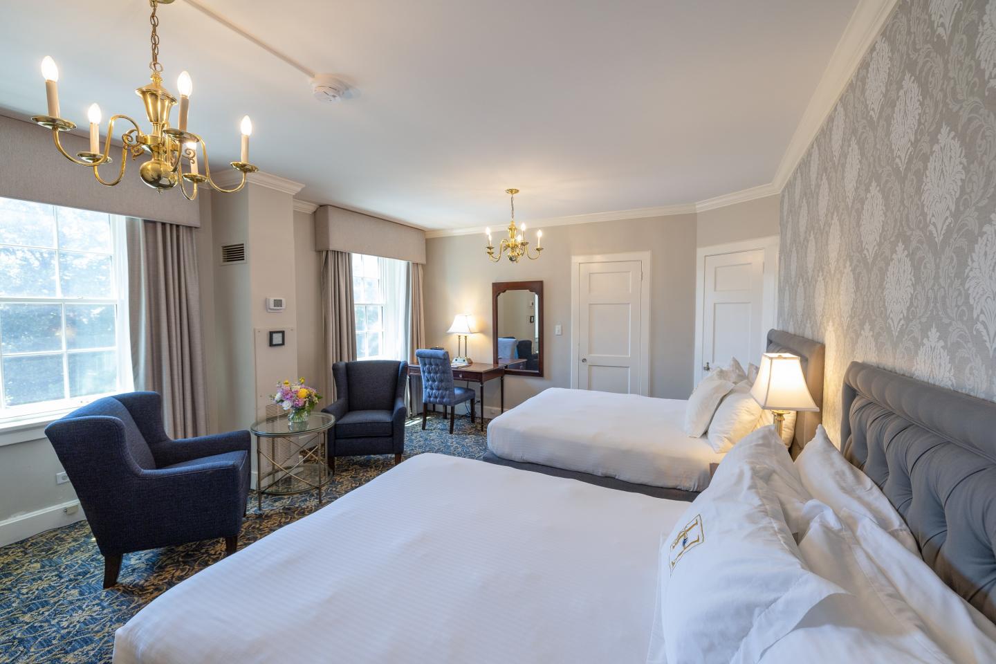 Elegant hotel room with two beds, armchairs, chandeliers, and large windows.