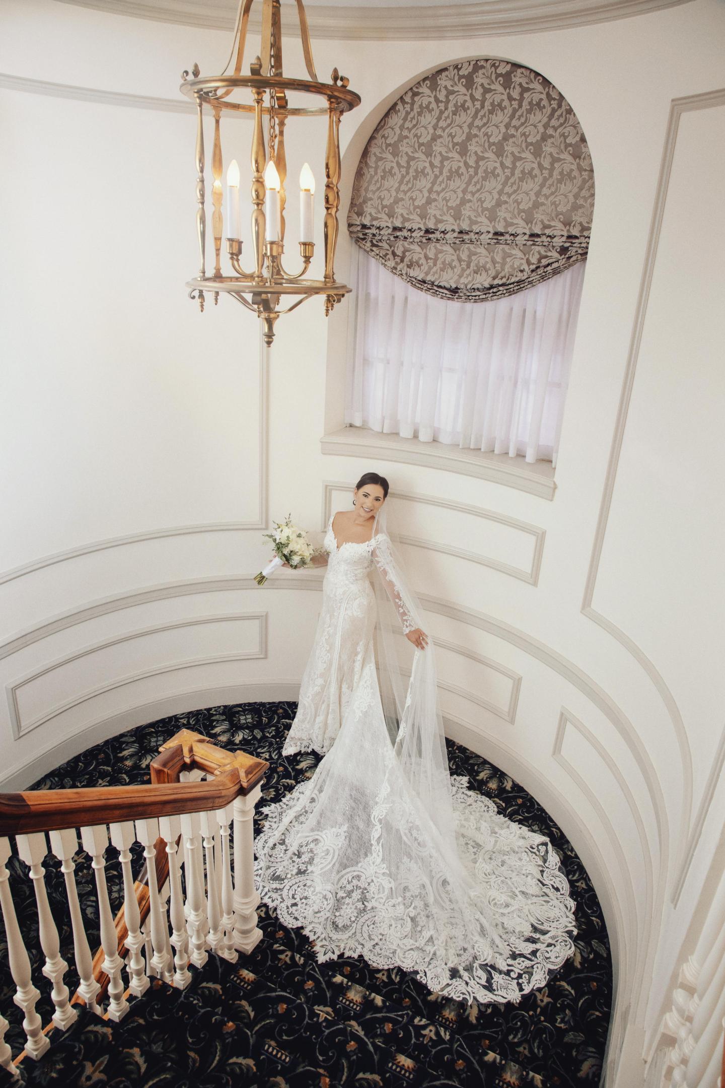 Bride in elegant white gown standing by a staircase with patterned carpet.