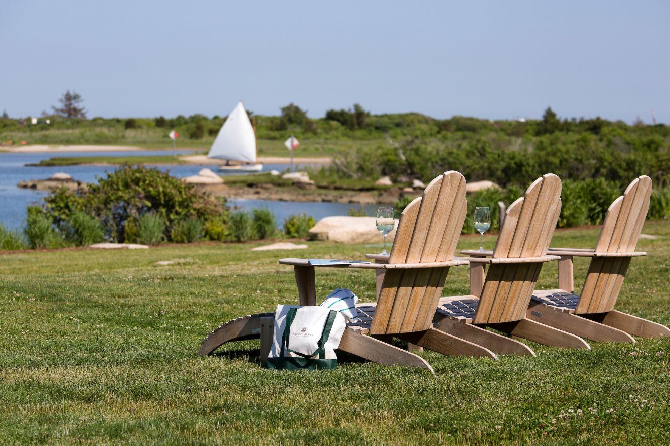 Wooden chairs on grass facing a lake with a white sail in the distan