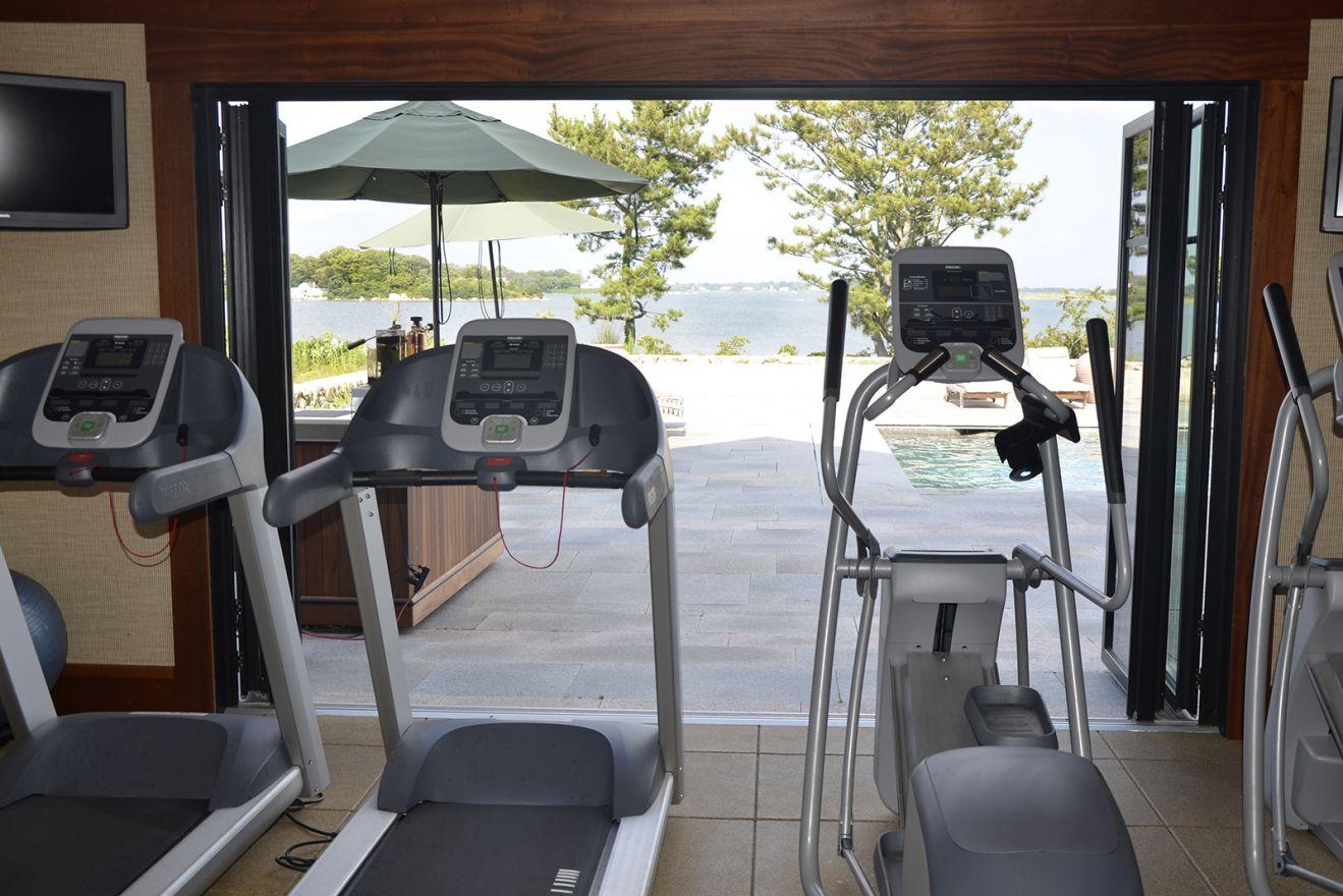 Treadmills and exercise bike face a patio with pool and trees, lake in the background.