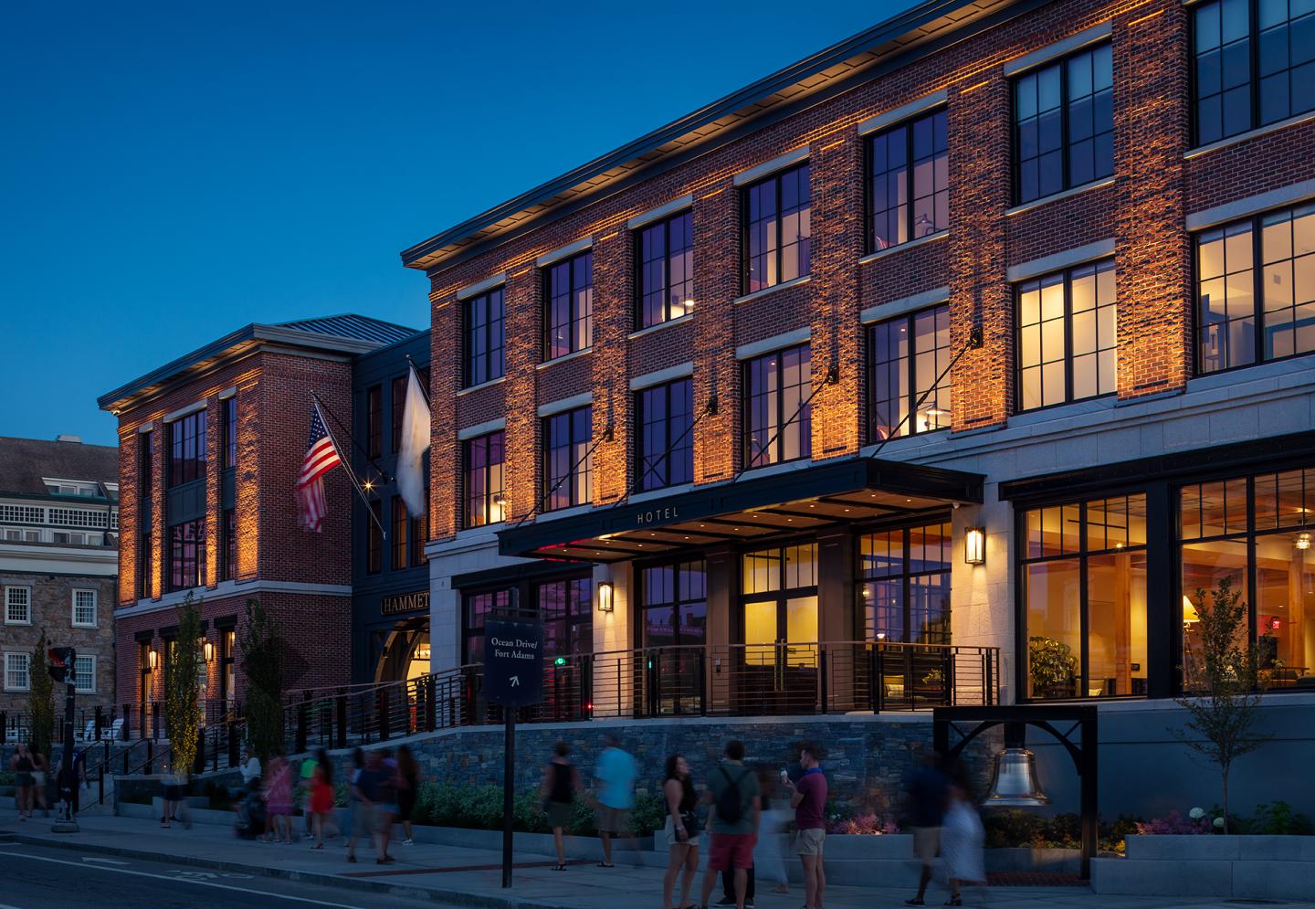 Brick building at dusk with lit windows and people walking outside.