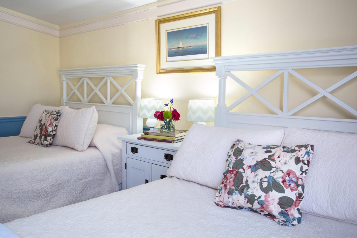 Two white beds with floral pillows in a cozy room, a painting on the wall above.