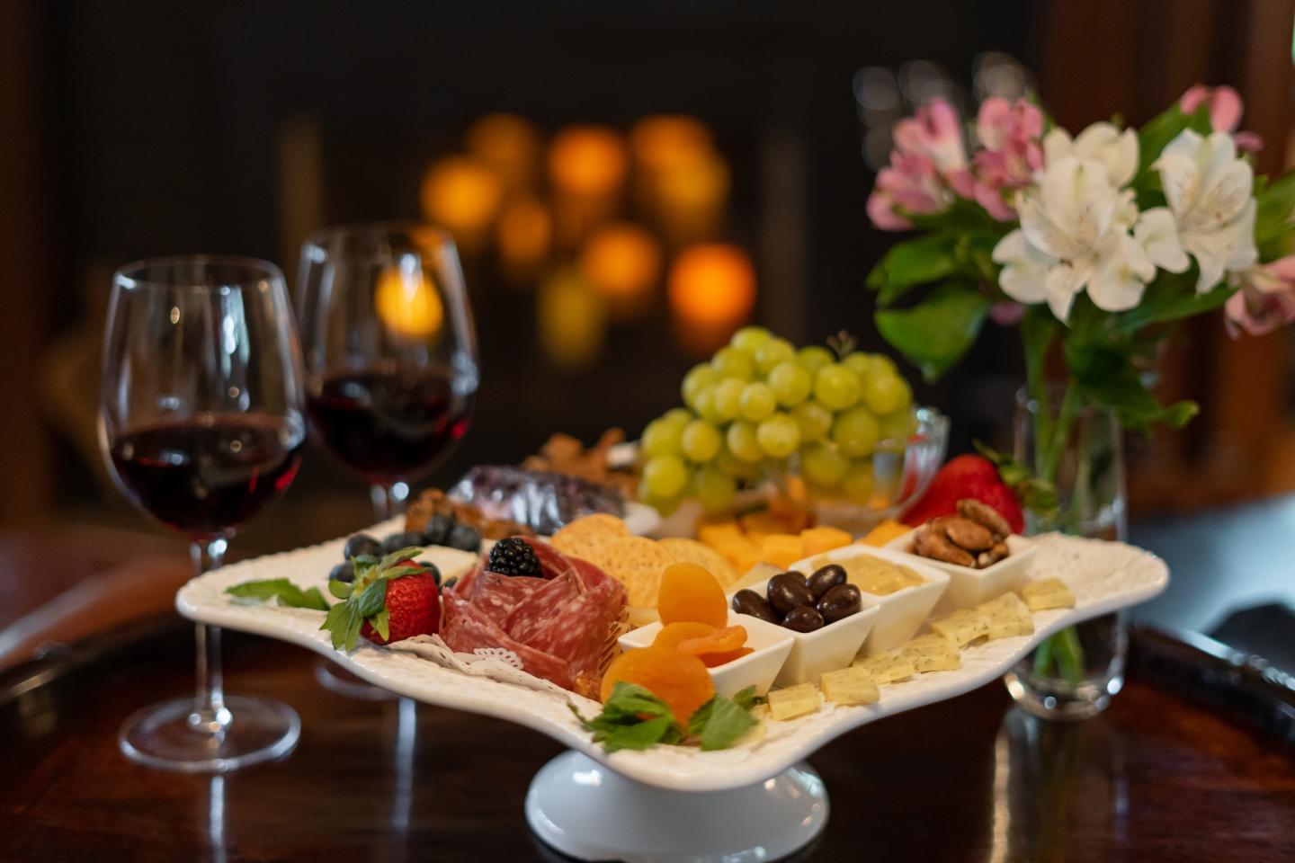 Cheese platter with fruits, two wine glasses, and flowers, set in a cozy fireside ambiance.