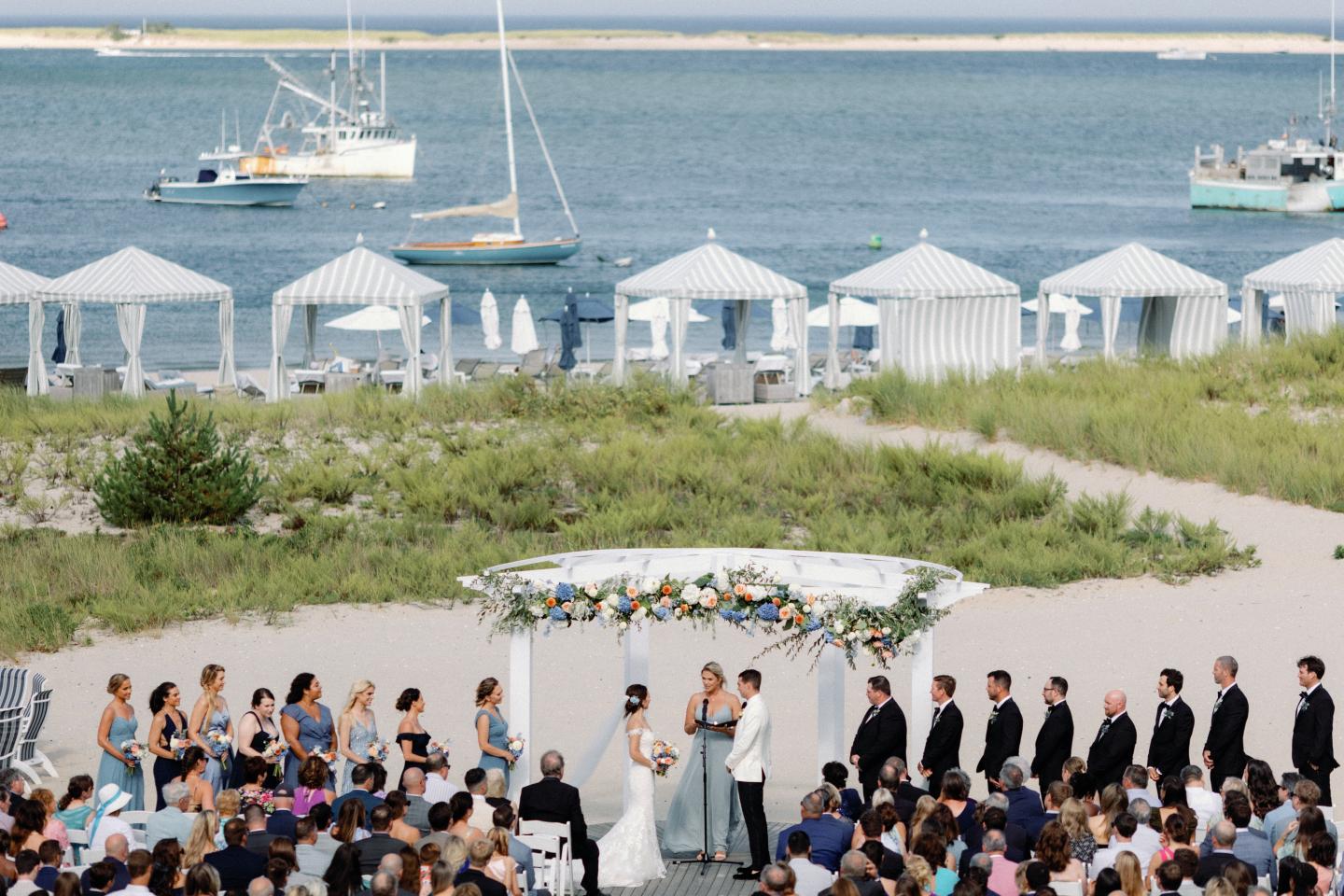 Beach wedding ceremony with guests seated, ocean in background.
