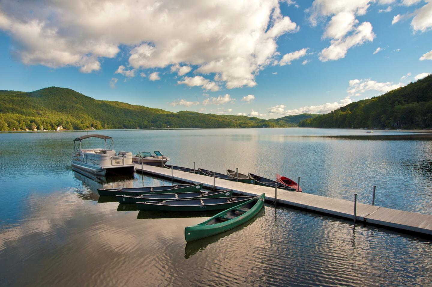 Dock with canoes and a boat on a tranquil lake, surrounded by lush green hills.