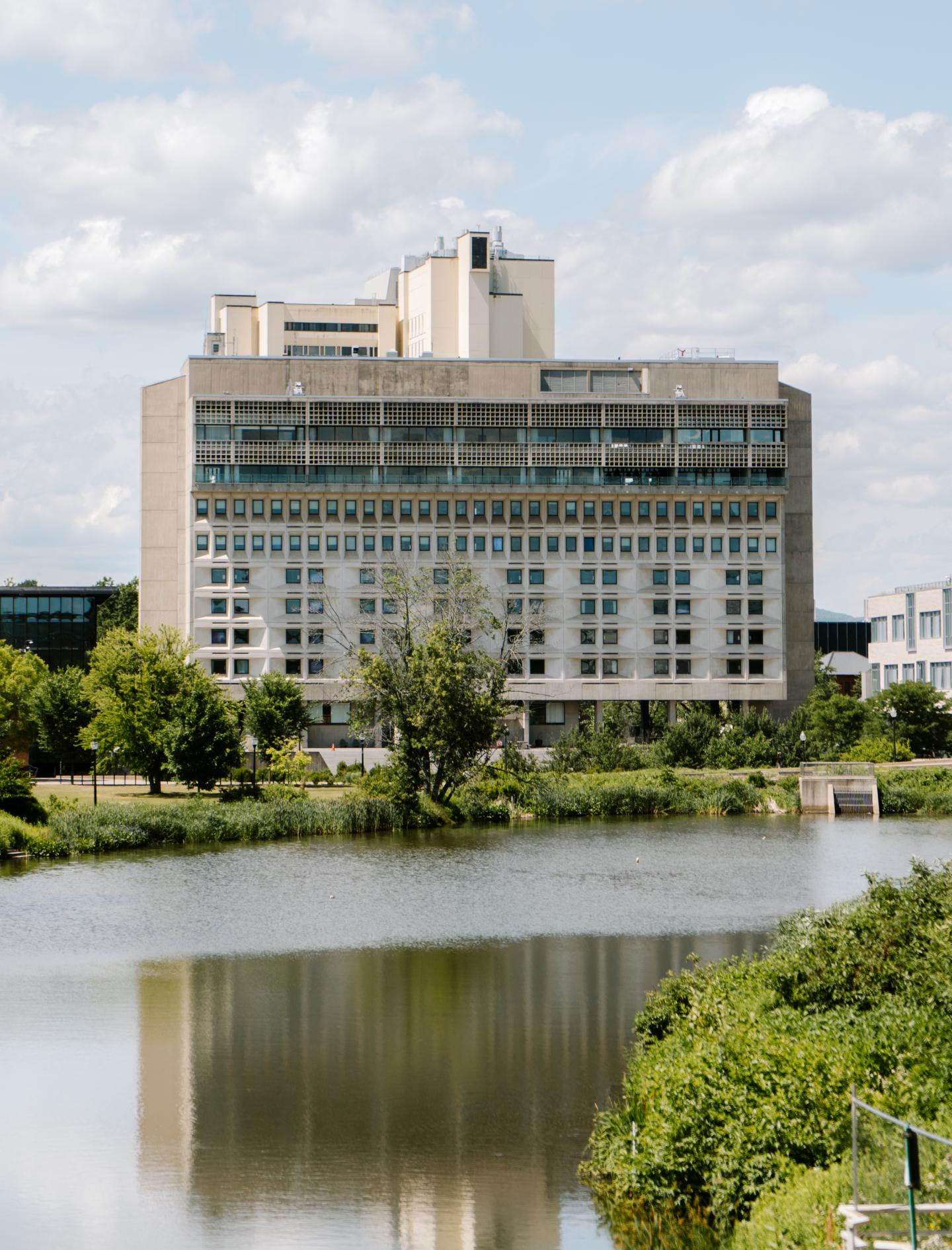 Tall building by a calm lake with greenery under a partly cloudy sky.