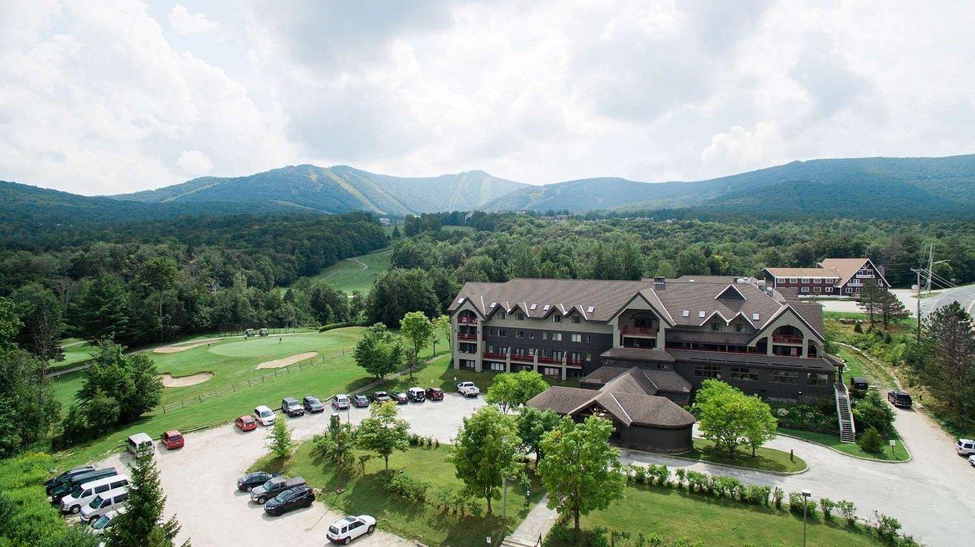 Aerial view of a lodge surrounded by trees, mountains, and a golf course under a cloudy sky. 04.2028