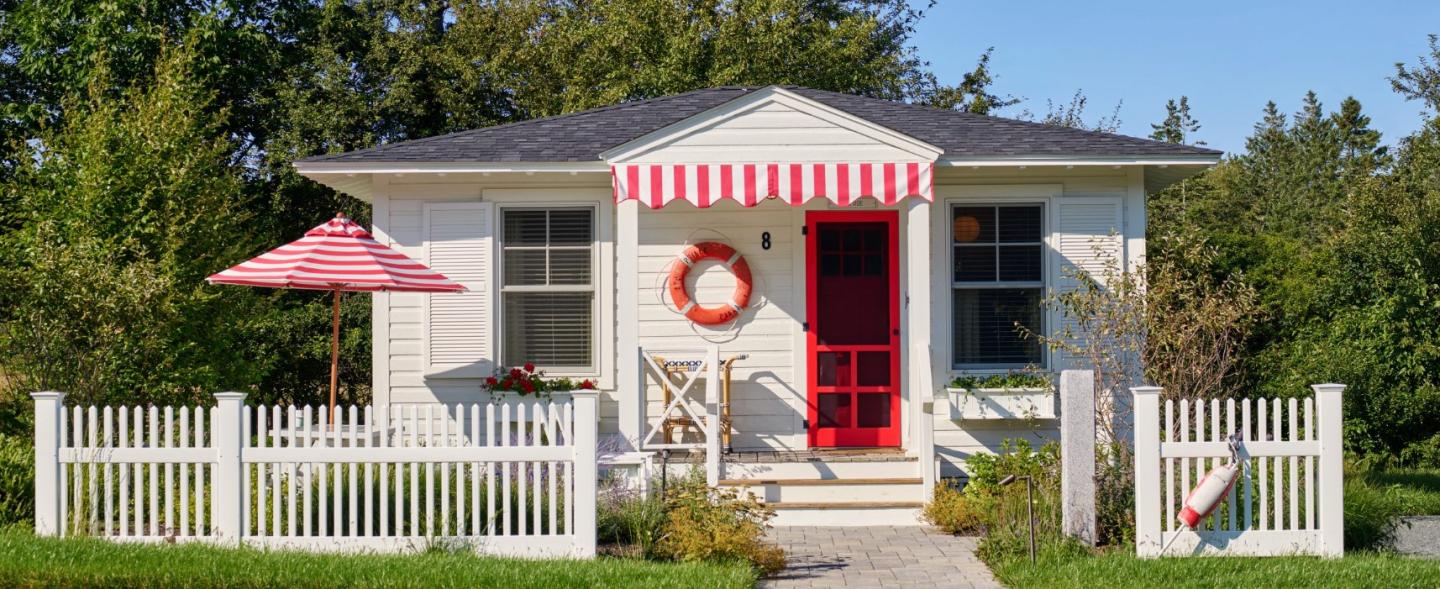 White cottage with red door, striped awning, and picket fence.