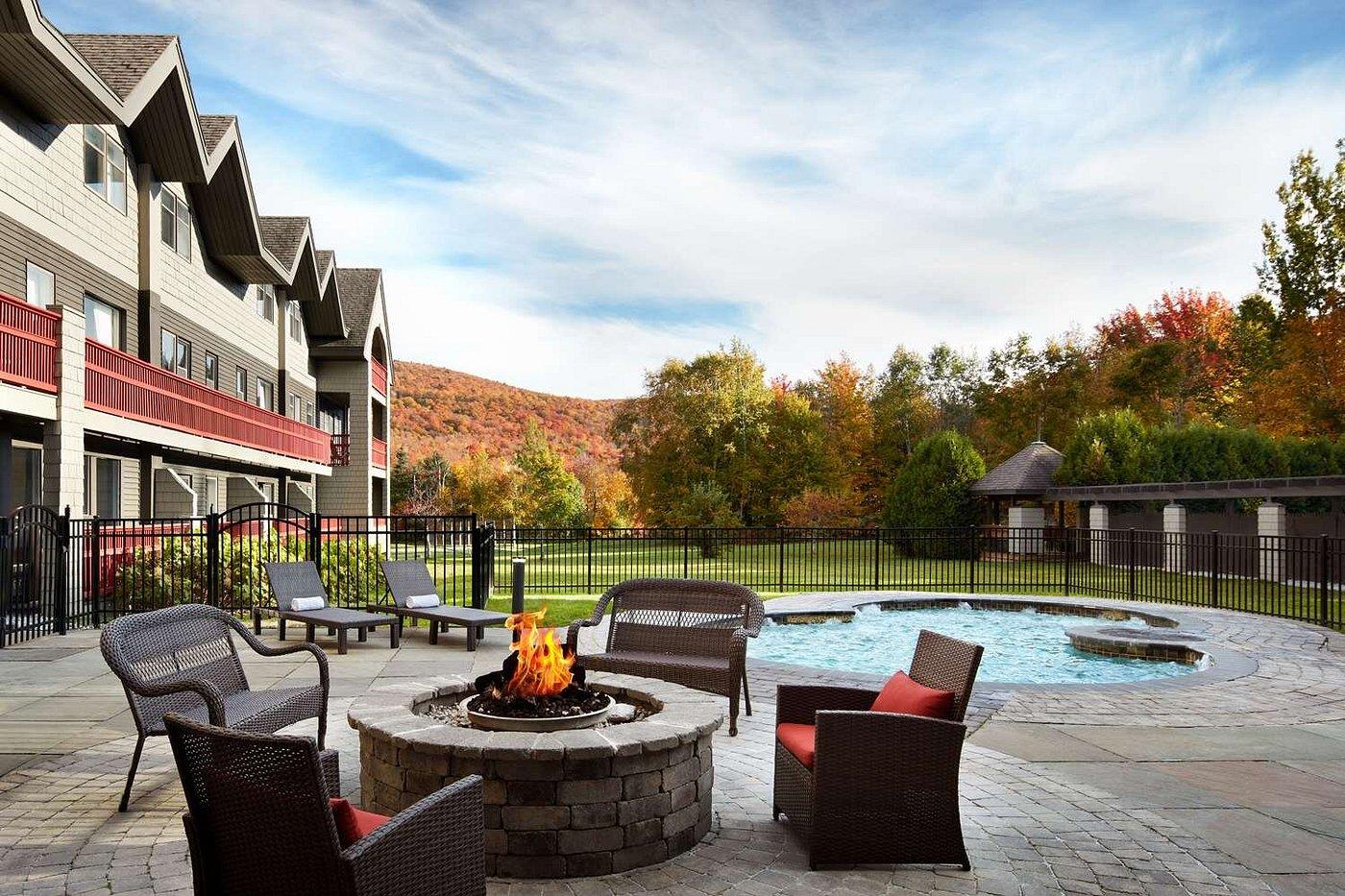 Firepit seating by a pool, autumn trees in background, large house adjacent.