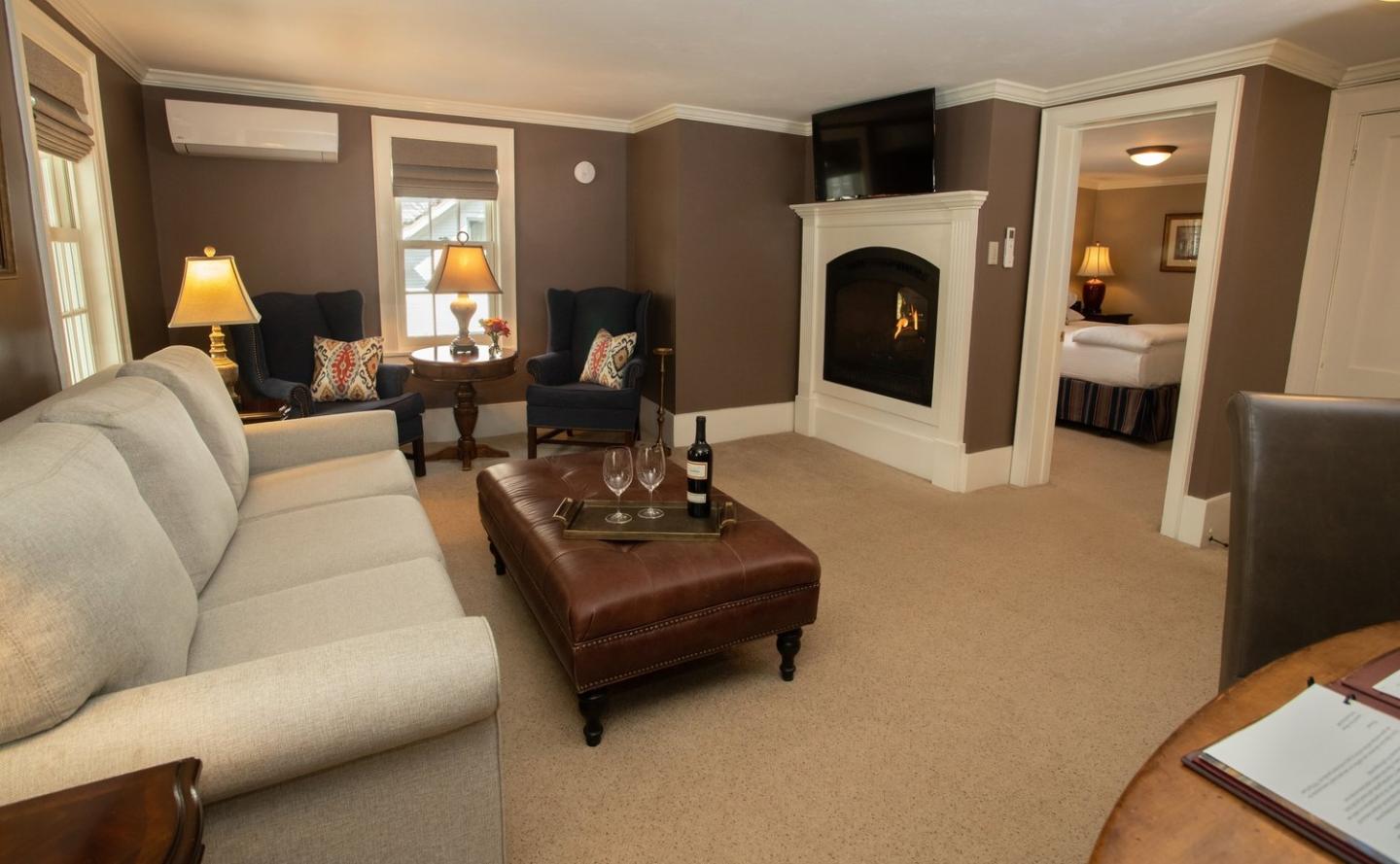 Cozy living room with beige sofa, armchairs, and fireplace. Bedroom visible through doorway.