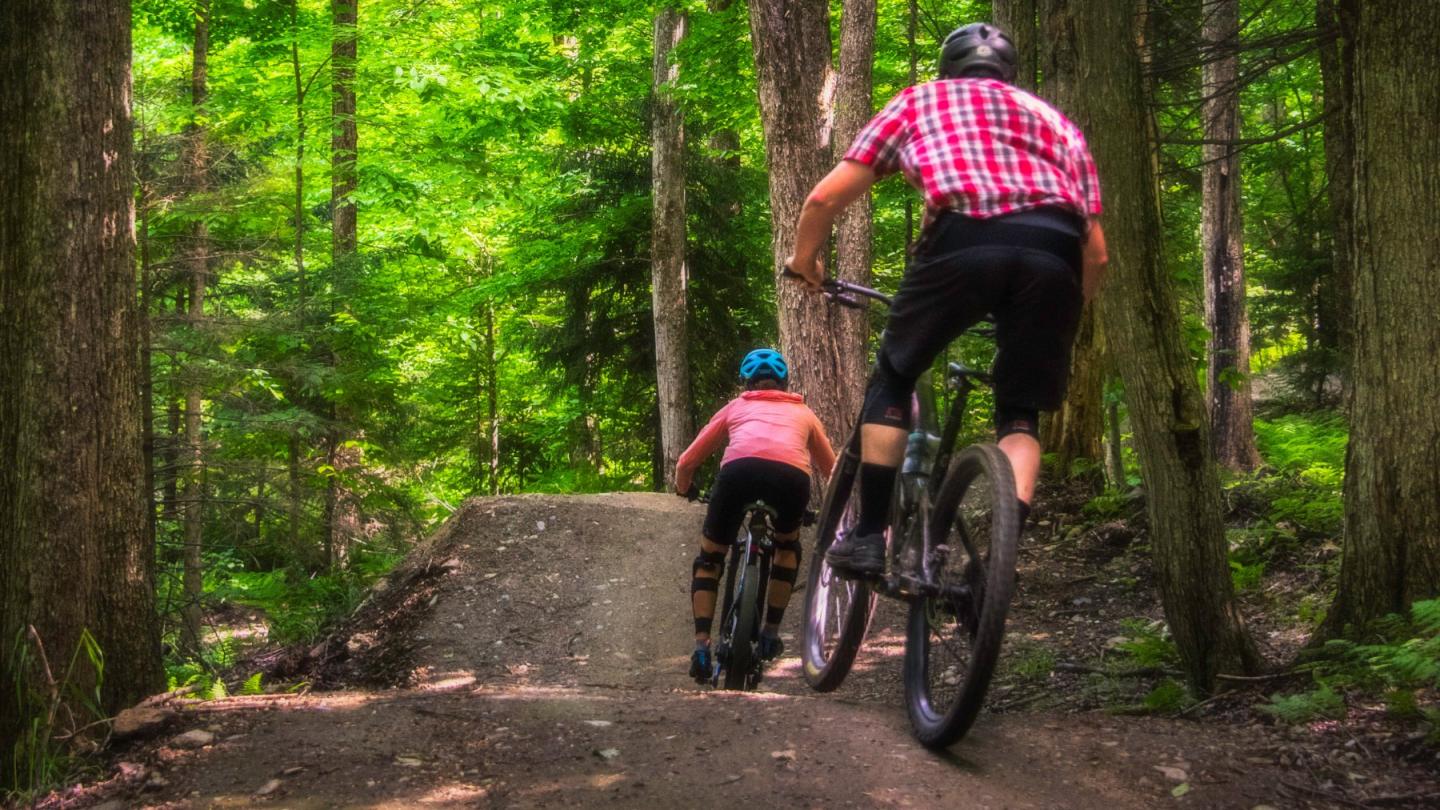 Two cyclists ride on a forest trail under lush green trees.
