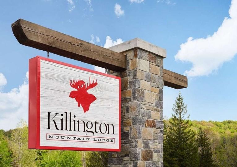 Stone pillar with a wooden sign, blue sky and trees in the background.