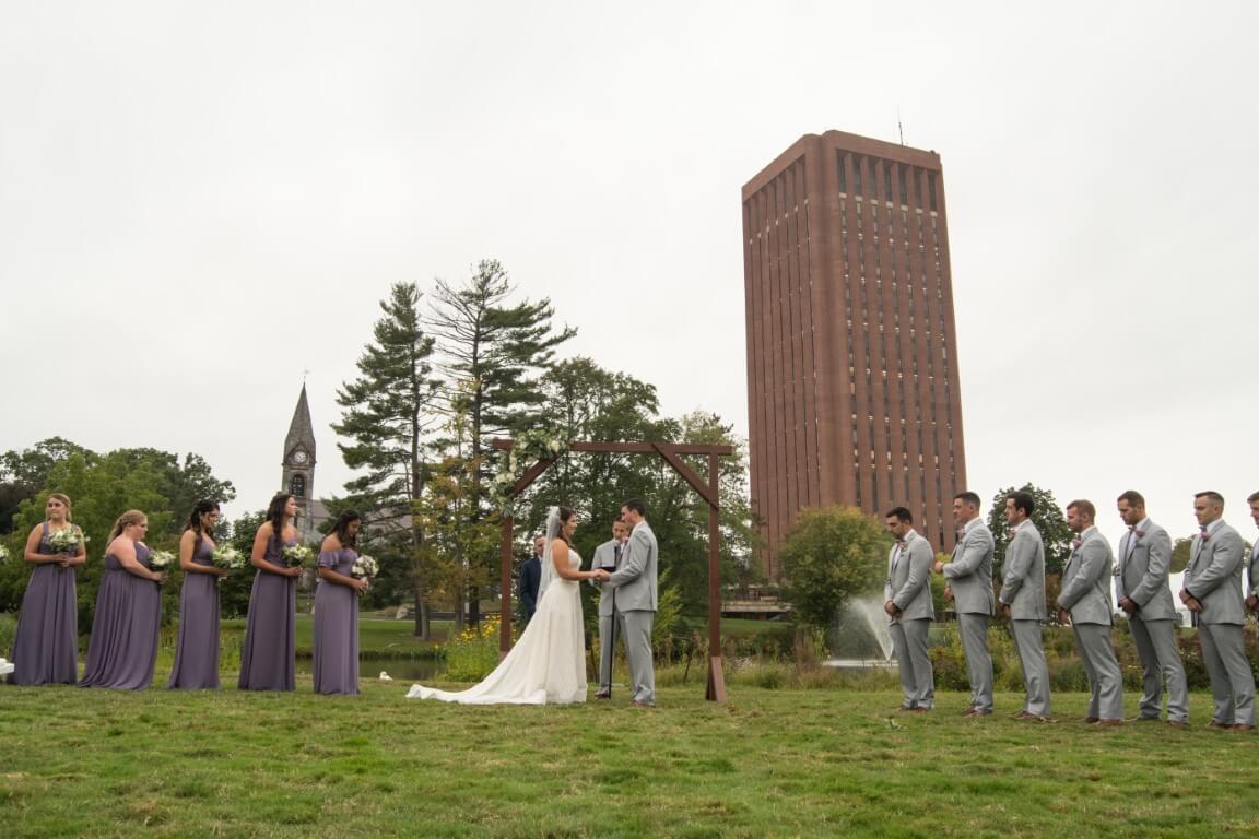 Wedding ceremony outdoors, couple under arch, bridesmaids in purple, groomsmen in gray.