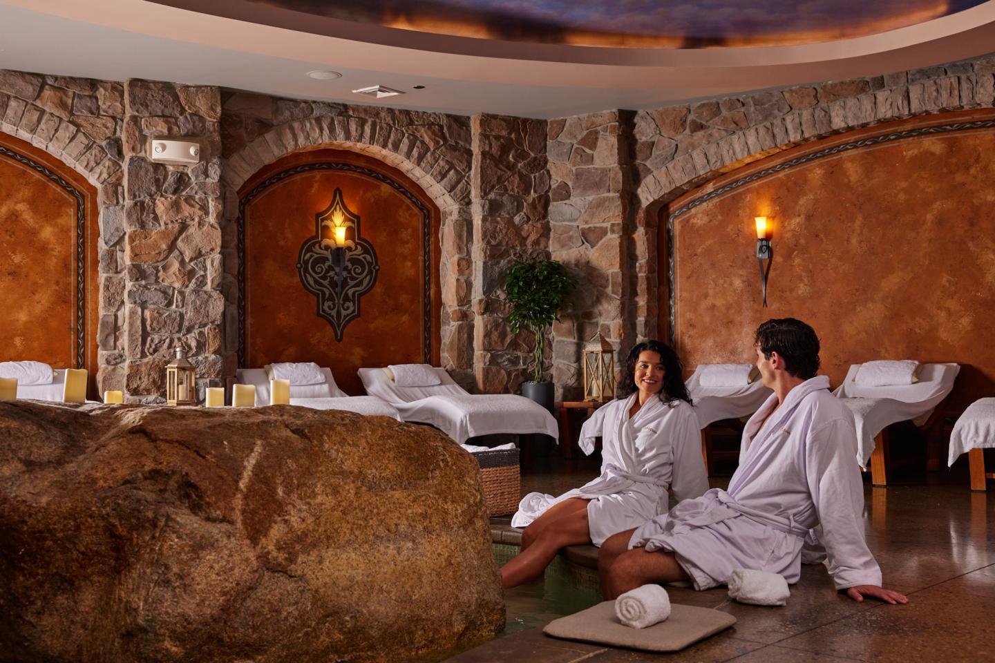 Cozy spa room with two people in robes seated on cushions, stone walls and warm lighting.