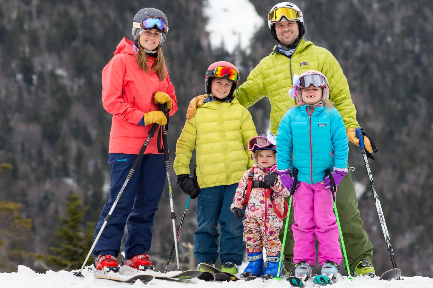 Family of five in colorful ski gear, smiling on snowy mountain.