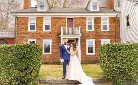 Bride and groom in front of a brick house, surrounded by green hedges.