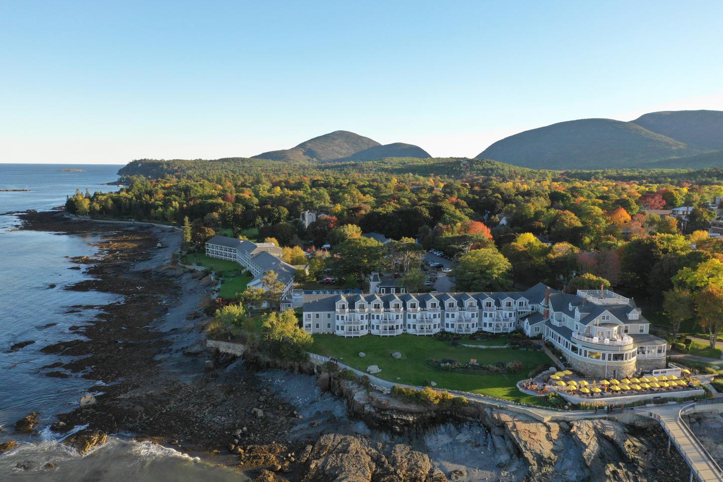 Coastal hotel surrounded by trees and mountains under clear sky.