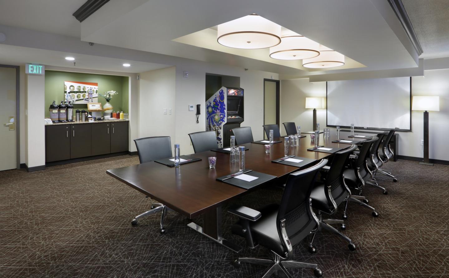 Modern conference room with a large table, chairs, and a refreshments area at the back.
