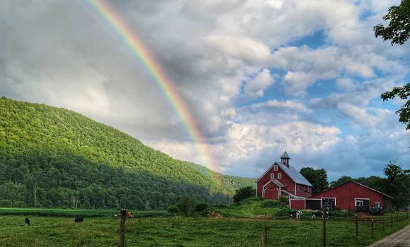 Rainbow over green hills and a red barn, under a partly cloudy blue sky.