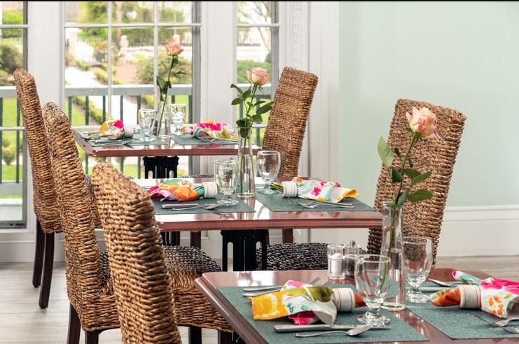 Dining tables with wicker chairs, set with colorful napkins and glassware, near large windows.