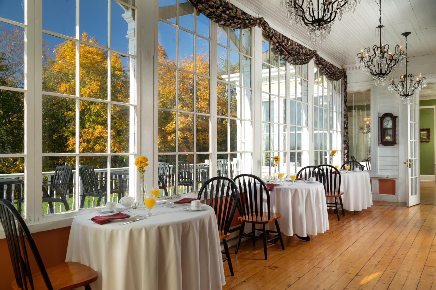 Sunlit solarium with white tablecloths, large windows, and autumn trees outside.