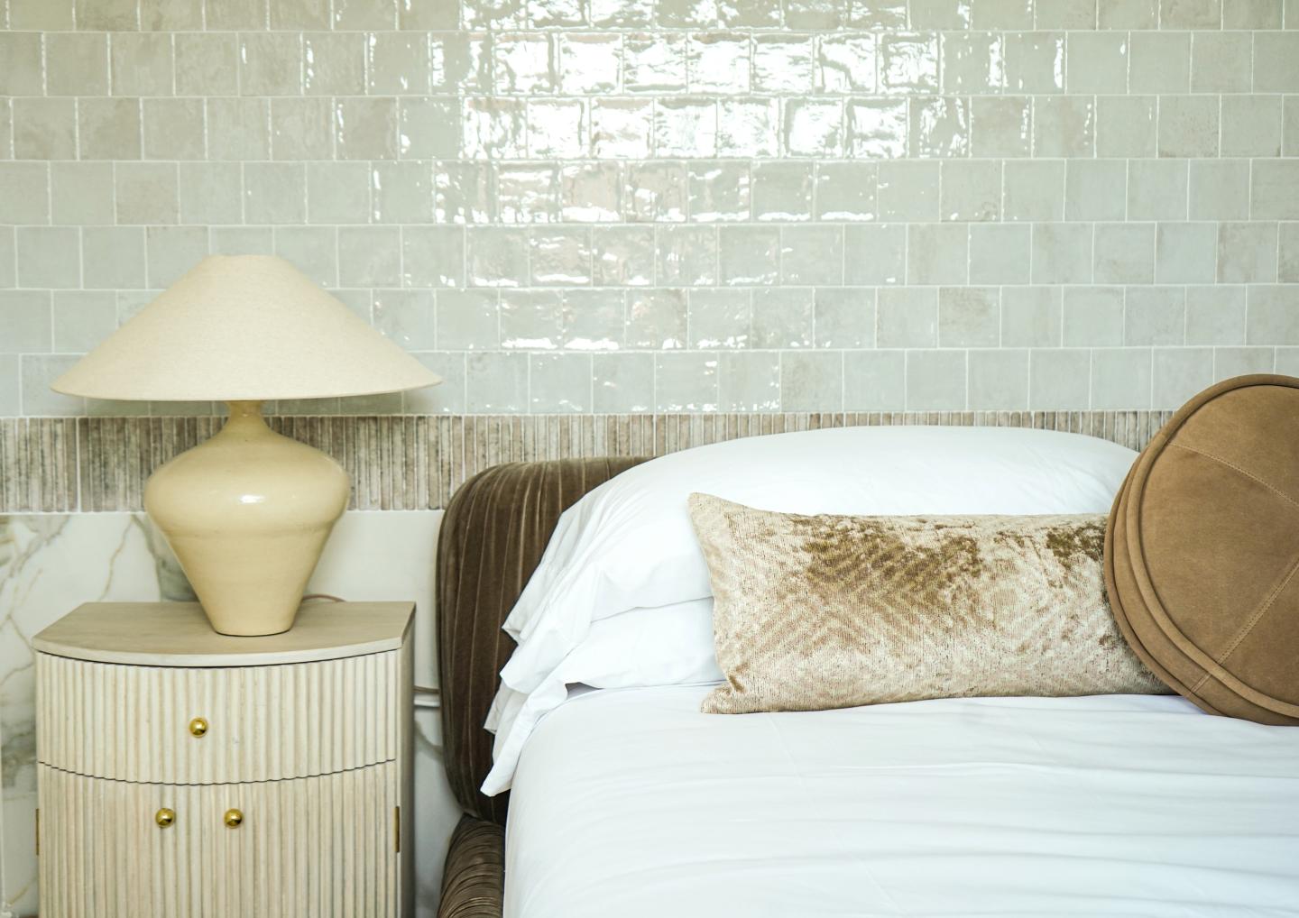Bed with white linens, beige pillows, and a lamp on a nightstand against a tiled wall.