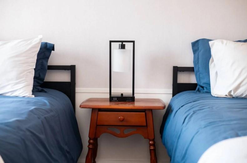 Two beds with blue bedding, a wooden nightstand, and a modern lamp between them.